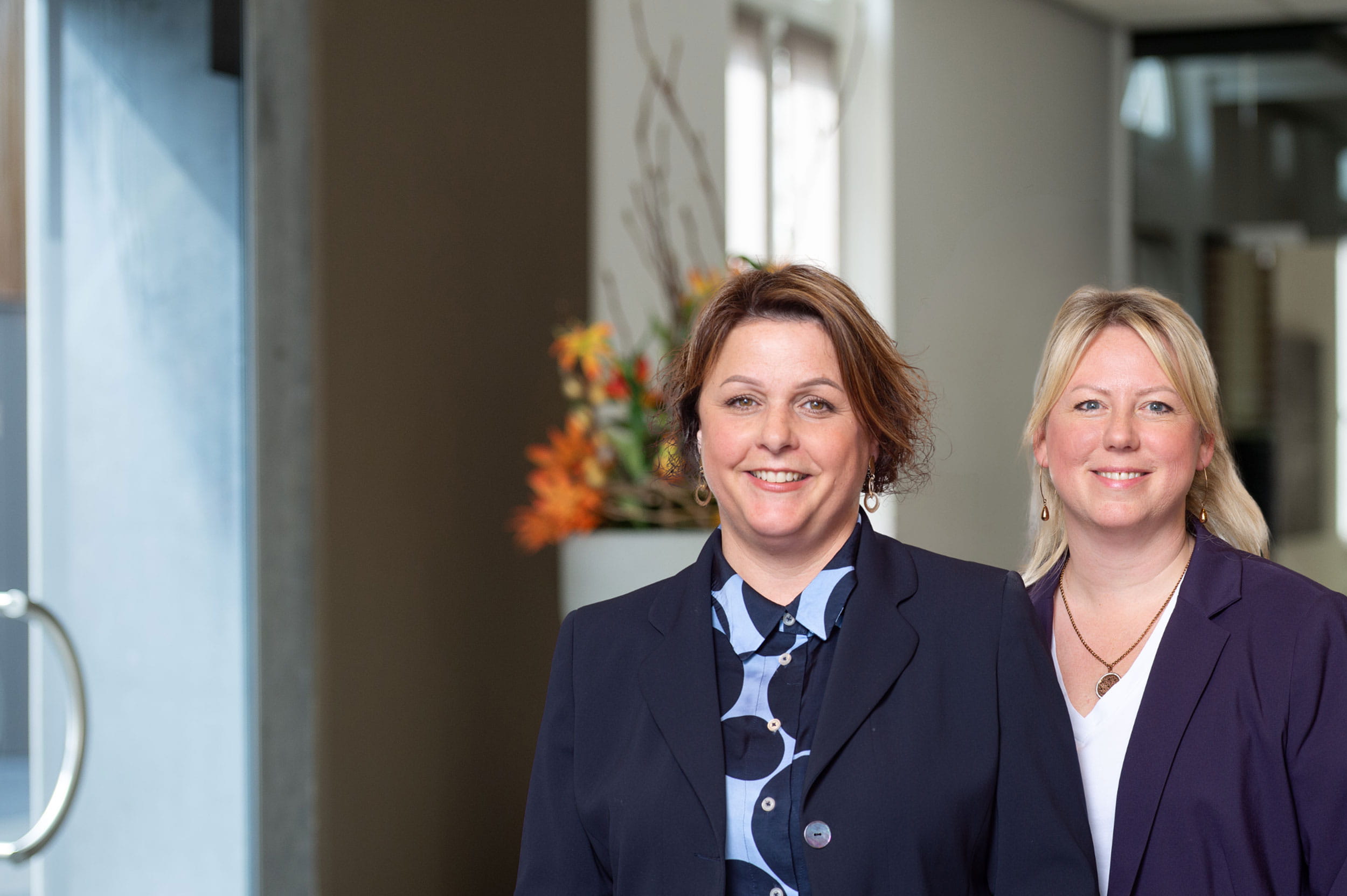 Twee personen in formele kleding staan naast elkaar binnen in een ruimte, met een glazen deur en een bloemenarrangement op de achtergrond.